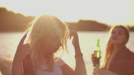 A-hot-young-blonde-beautifully-moves-her-hands-in-dance-on-the-hot-party-on-the-beach-with-beer.-This-is-a-happy-summertime-on-the-open-air-party-on-the-sand-beach-near-the-lake.